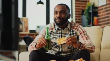 Neşeli genç adam bira içiyor ve pizza yiyor, evde kanal programında film izliyor. Fast food yemek ve alkolle yemek yiyip film izleyen mutlu bir adam. El kamerasıyla..
