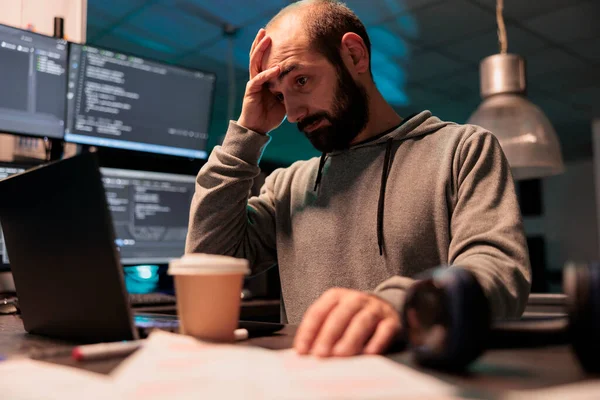 Stressed overworked developer programming html code on laptop and multiple monitors, working under stress after hours. Male app programmer feeling frustrated and tired coding data script.