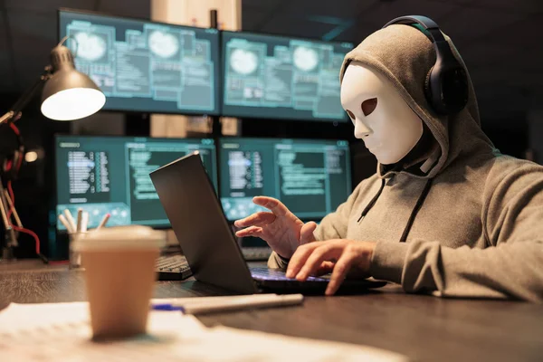 Hacker with mask and hood breaking security server, installing virus to create computer malware and steal big online data. Masked criminal scammer hacking netowrk system at night.