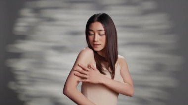 Asian woman expressing tenderness and confidence in new skincare ad campaign, using natural cosmetics in studio. Flawless cheerful girl feeling sensual and elegant on camera, self love.