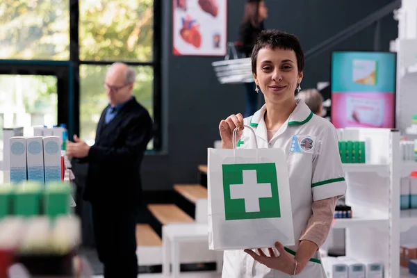 Pharmaceutical employee holding medical retail store shopping bag, smiling and looking at camera. Young drugstore worker standing in apothecary aisle and showing package with medicaments portrait
