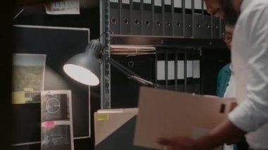Detective partners discussing investigation and clues found in records folders, private felony case in police archive room full of racks. Inspectors team studying evidence board. Handheld shot.