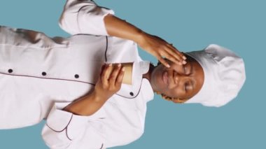 Vertical video: Exhausted chef yawning and drinking coffee on camera, trying to not fall asleep at culinary service job. Woman cook with apron enjoying cup of caffeine refreshment over blue background
