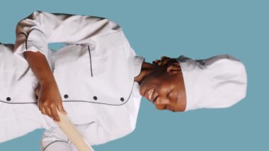 Vertical video: Smiling positive cook playing with rolling pin and singing on camera, having fun with cooking utensils. Young female chef holding wooden paddle, gastronomy tools over blue background.