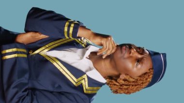 Vertical video: Young aircrew member thinking about new ideas, brainstorming solutions on camera. Professional stewardess in aviation flying uniform, feeling pensive on blue background. Portrait.