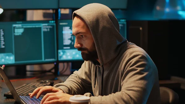 IT fraud criminal breaking through cyber security system and stealing data on multiple monitors. Dangerous hacker with hood drinking coffee and causing computer server malware with virus.