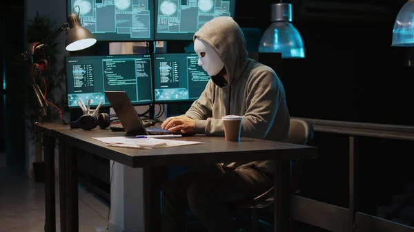 Masked person hacking computer network at night, working on laptop and multiple monitors to steal IT information. Hacker with hidden identity acting dangerous, server security breach. Handheld shot.