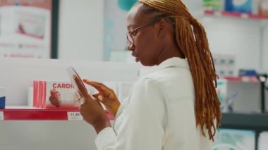 Afro-Amerikalı kadın eczanede ilaç paketi okuyor, sağlık ürünlerine bakıyor. Eczacılık, uyuşturucu ve ilaç kutularını kontrol eden genç bir yetişkin. El kamerasıyla..