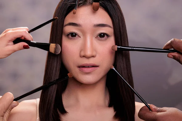 Fashion model getting make up in professional studio and looking at camera. Makeup artists team hands holding brushes around young adult attractive asian woman close up portrait