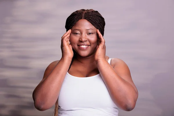 African american body positive model touching fresh healthy skin and looking at camera with happy facial expression. Beautiful plus size woman posing in studio for skincare cosmetology