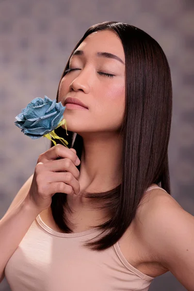 Asian woman with closed eyes enjoying blue rose smell. Elegant young beauty model with fresh glowing skin and smooth brown hair holding flower in hand, wearing casual beige top