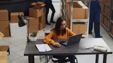 Young employee with impairment working in warehouse, doing desk work for stock logistics and inventory on laptop. Woman wheelchair user doing financial planning in disability friendly space.