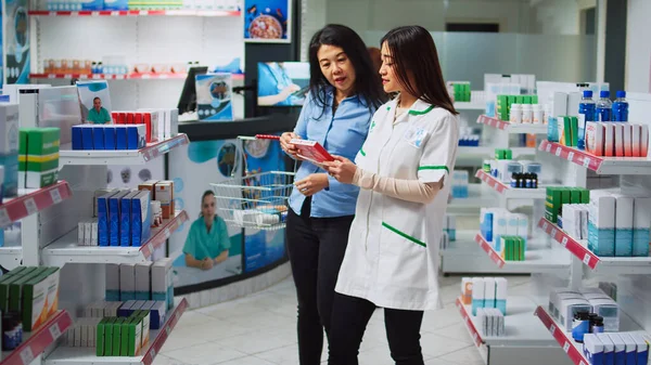 若い薬剤師はドラッグストアの女性を支援し アジアの顧客に薬や薬を与えるために処方紙を読んでいます 薬や薬の箱の話をしている人たちは — ストック写真