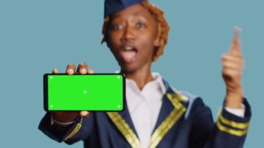 Kadın uçak mürettebatı kameradaki mavi arkaplanda yeşil ekran görüntüsünü işaret ediyor. İzole edilmiş telif alanı ve havacılık üniforması giymiş krom anahtar şablonu gösteren genç hostes.