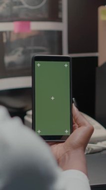 Dikey video: Özel dedektif yeşil ekranlı akıllı telefonu tutuyor, polis memuru izole edilmiş bir telif alanı görüntüsüne bakıyor. Ofisteki krom anahtar modelleme şablonu kullanılarak kanıt odasındaki araştırmacı.