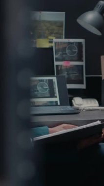 Dikey video: Özel dedektif polis arşiv odasında suç bilgi kayıtlarını okuyor, bilgisayardaki gözetim fotoğraflarını kontrol ediyor. Güvenlik ajanı ofiste dava dosyasını inceliyor.