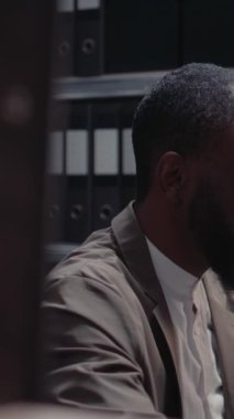 Dikey video: Afro-Amerikan ajanı dosyalarla suç davasında çalışıyor, adli kanıtları ve ofis alanındaki belgeleri inceliyor. Profesyonel soruşturma yürüten özel dedektif. Üçayak atışı