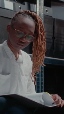 Dikey video: Afro-Amerikan ajanı gözetim fotoğraflarını analiz ediyor, şüphelileri yakalamak için önemli polis dosyalarına bakıyor. Kolluk kuvvetleri dedektifi adli tıp uzmanlık raporunu tutuyor. El bilgisayarı