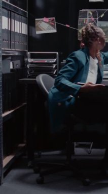 Dikey video: Afro-Amerikan kadın polis dava dosyalarını inceliyor, adli kanıtlarla ve şüpheli fotoğraflarla adli soruşturma üzerinde çalışıyor. Kadın müfettiş, hukuk bürosunda detektif kurulunu kullanıyor..