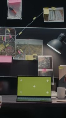 Dikey video: Polis ajanları dizüstü bilgisayardan yeşil ekrana bakıyor, polis arşivlerindeki suç kayıtlarını inceliyor. Özel dedektifler boş kroma anahtarıyla izole edilmiş model şablonu inceliyor..