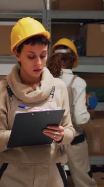 Dikey video: Kargo envanteri ve lojistik yapmak için dosyaları kullanan kadın çalışan kağıtlardaki mal listesini kontrol ediyor. Tulumlu kadın depodaki karton kutu ya da paket sayısını sayıyor..