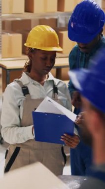 Dikey video: Depo çalışanları dizüstü bilgisayarda sevkiyat planlıyorlar, depo deposunda kutularda mallarla çalışıyorlar. Afro-Amerikalı insanlar hisse senedi lojistiklerini iş dağıtımı için kontrol ediyorlar.