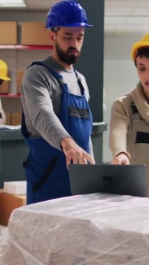 Dikey video: Erkek ve kadın eşyaları kutulara koymayı, dizüstü bilgisayarlardaki hisse senedi lojistiğine bakmayı ve ürünleri saymayı düşünüyor. Depo çalışanı ve amirinin tulumu ve şapkası var.