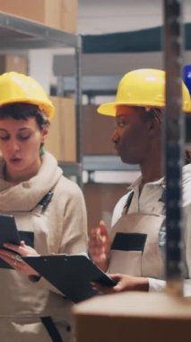 Dikey video: Çok ırklı denetim ekibi ambar raflarına konulan malları sayıyor, paketlerdeki ürünlere bakıyor. Depoda çalışan işçi tulumlu genç kadınlar..