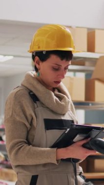 Dikey video: Depo deposunda tarayıcı ve tablet kullanan kadın denetleyici ürünleri kutularda tarıyor. Genç yetişkin işçilerle lojistik ve planlama siparişi nakliyatı üzerinde çalışıyor..