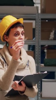 Dikey video: Depo denetleyicisi kargo envanteri yapmak için kağıtları kullanarak dosyalardaki mal listelerini kontrol ediyor. Tulumlu ve kasklı bir kadın depo raflarındaki kutu ve paket sayısını sayıyor..