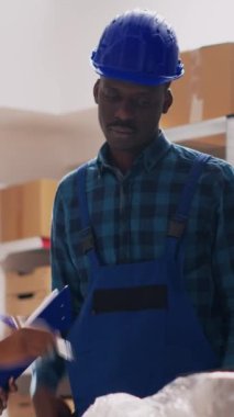 Dikey video: Depoda kalite kontrol yapan kadın denetçi, depo raflarından stok ürünleriyle çalışıyor. Çalışanlardan oluşan bir ekip teslimat için malları kontrol ediyor. El kamerasıyla..