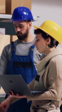 Dikey video: Çeşitli çalışanlar depodaki stok lojistiği hakkında konuşuyor, depodaki raflara konan mallar. Depoda dizüstü bilgisayar kullanan kadın ve erkek tulum giyiyor.