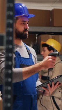 Dikey video: Depodaki paketlerdeki barkodları tarayan erkek denetçi, tarayıcıyı karton kutulara doğrultuyor ve stokları kontrol ediyor. Tulumlu bir adam depoda tablet tutuyor, endüstriyel mallar..