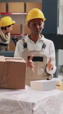 Dikey video: Afro-Amerikan kadın reklam ürünleri kutularda depolama odasında akıllı telefonla çalışıyor. Tulum giymiş kadın çalışan sosyal medya ve endüstriyel stoklar için reklam videosu çekiyor.