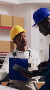 Dikey video: Afro-amerikan insanlar depoda kalite kontrol yapıyorlar, depo raflarındaki ürünlerle çalışıyorlar. Erkek ve kadından oluşan bir ekip endüstriyel hizmet için ürünleri kontrol ediyor. El kamerasıyla..