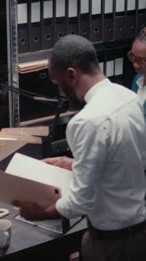 Dikey video: İki polis memuru arşiv odasındaki güvenlik kamerası görüntülerinden bahsediyor, detayları ortaya çıkarmak için suç davasını inceliyorlar. Dedektifler dedektiflik yapıyor, soruşturma yürütüyor.