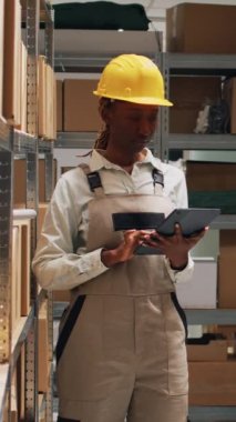 Dikey video: Tulum giymiş kadın çalışan tablet envanterini kontrol ediyor, malları nakletmek için lojistik planlaması yapıyor. Genç yetişkin sanayi stoklarını kutular, paket dağıtımı ve