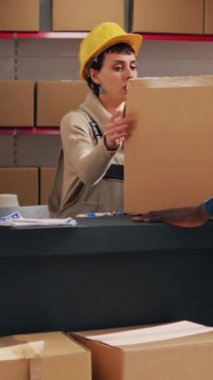 Dikey video: Kadın denetçi depoda barkodlu ürünleri tarıyor, karton paketlerde endüstriyel dağıtım planlıyor. Ürün kutuları ve dijital tablet üzerinde tarayıcı kullanan genç kadın.