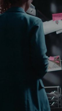 Dikey video: Dedektif arşivde suç soruşturması yürütüyor ipucu ve fotoğraflarla dolu kanıt haritasına bakıyor. Kadın Umman gözetim bilgilerini ve adli kayıtları inceliyor.