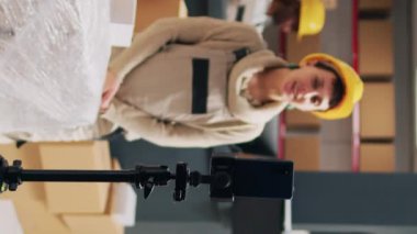 Dikey video: Denetmen depoda reklam videosu kaydediyor, ticari paketleri sosyal medya uygulamasında gösteriyor. Depo çalışanı halkla ilişkiler reklamı ve depo rafları için akıllı telefon kullanıyor..