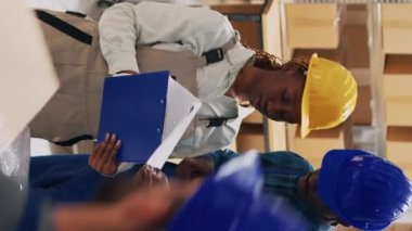 Dikey video: Depo çalışanları dizüstü bilgisayarda sevkiyat planlıyorlar, depo deposunda kutularda mallarla çalışıyorlar. Afro-Amerikalı insanlar hisse senedi lojistiklerini iş dağıtımı için kontrol ediyorlar.