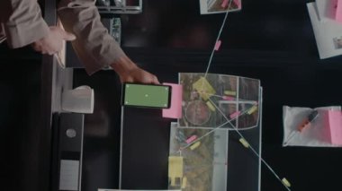 Dikey video: Polis ortakları olay odasında izole şablon kullanarak akıllı telefonu yeşil ekranla analiz ediyorlar. İki özel dedektif krom anahtarlı cep telefonu görüntülerine bakıyor..