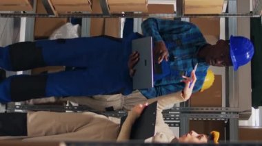 Dikey video: Çok ırklı çalışanlardan oluşan bir ekip dizüstü bilgisayarda stok envanterini kontrol ediyor, depoda kargo bulunuyor. Dağıtımı planlamak için endüstriyel malları analiz eden depo işçileri ve