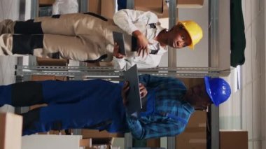 Dikey video: Afro-amerikan insanlar dizüstü bilgisayar ve tablet üzerindeki malları analiz ediyorlar, depolama alanında stok envanteri oluşturmak için cihazlar kullanıyorlar. Karton paketler ve kargolarla çalışan genç adam ve kadın.