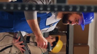 Dikey video: Depodaki paketlerdeki barkodları tarayan erkek denetçi, tarayıcıyı karton kutulara doğrultuyor ve stokları kontrol ediyor. Tulumlu bir adam depoda tablet tutuyor, endüstriyel mallar..