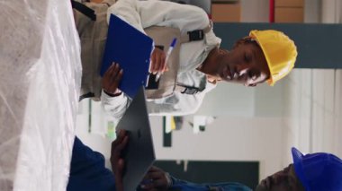 Dikey video: Tulum giymiş Afro-Amerikalı insanlar depo raflarına yerleştirilen ürünleri analiz ediyorlar, kargo sevkiyatlarını karton kutulara koyuyorlar. Endüstriyel ürünlerle çalışan kadın ve erkek, depo alanı.