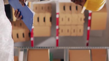Dikey video: Dosyalardaki ürünlerin listesini inceleyen çok ırklı bir ekip, envanter stokları hakkında konuşuyor. Tulumlu kadın ve erkek depodaki malları kontrol ediyor.