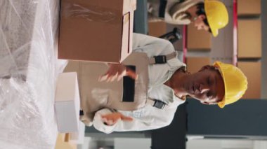 Dikey video: Afro-Amerikan kadın reklam ürünleri kutularda depolama odasında akıllı telefonla çalışıyor. Tulum giymiş kadın çalışan sosyal medya ve endüstriyel stoklar için reklam videosu çekiyor.