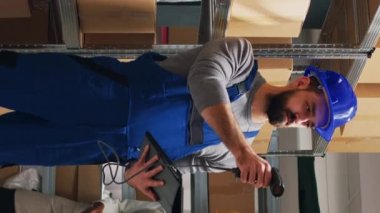 Dikey video: Depo çalışanı kutulardaki barkodları tarıyor, dağıtım ya da sevkiyat için kargoyu kontrol ediyor. Depoda tarayıcı ve dijital tabletle çalışan genç adam, endüstriyel ürünleri naklediyor..