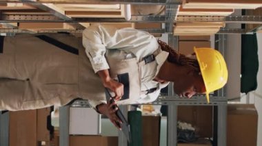 Dikey video: Tulumlu Afro-Amerikalı kadın tabletle çalışıyor, depo malları için stok lojistiği planlamak için cihaz kullanıyor. Depo çalışanı karton kutulardaki endüstriyel kargoyu kontrol ediyor..